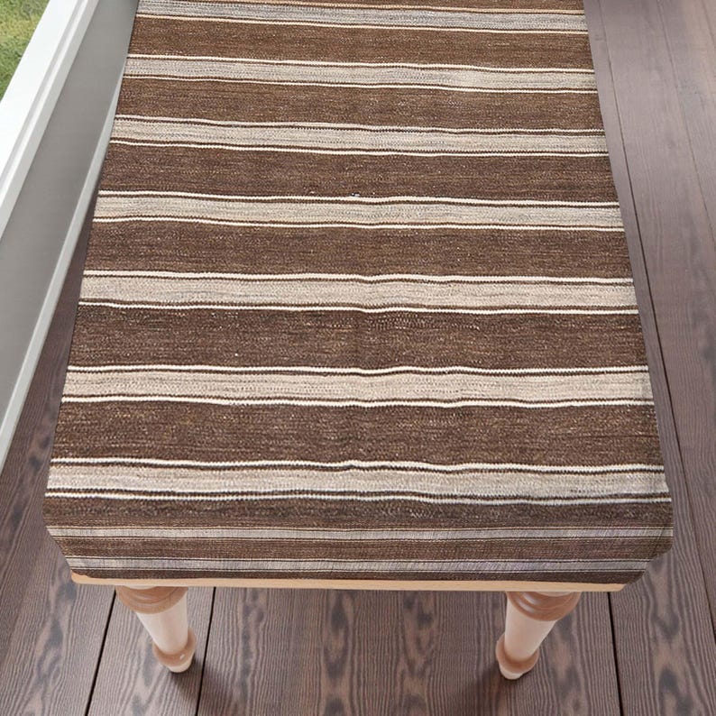 May include: A rectangular table runner with brown and beige horizontal stripes. The runner is placed on a wooden table with light-coloured legs. The design features stripes of varying widths and shades of brown and beige.