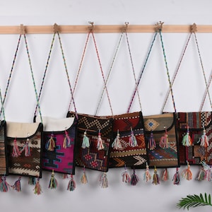 May include: Seven colorful patterned crossbody bags with tassels hanging from a wooden rack. The bags are made of woven fabric and have a black trim.