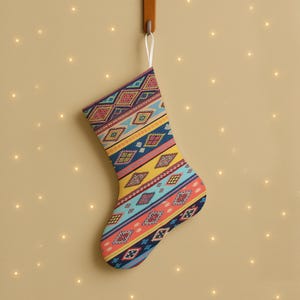 Könnte beinhalten: Ein Weihnachtsstrumpf mit einem farbenfrohen geometrischen Muster in Blau, Gelb, Rot und Orange hängt an einem Holzhaken. Der Strumpf hat eine weiße Schlaufe zum Aufhängen und befindet sich vor einem beigefarbenen Hintergrund.
