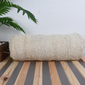 May include: A cream-colored, rolled up, woven throw pillow on a wooden slatted surface. A green potted plant is in the background.
