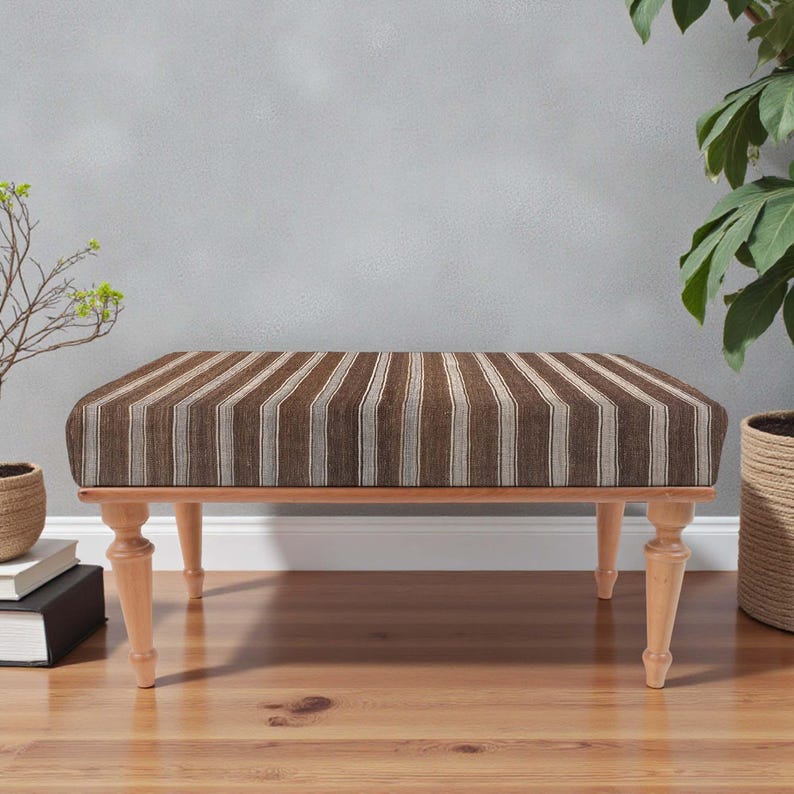 May include: A rectangular bench with a brown and beige striped pattern. The bench has four light-coloured wooden legs and a wooden frame. The bench is placed on a wooden floor.