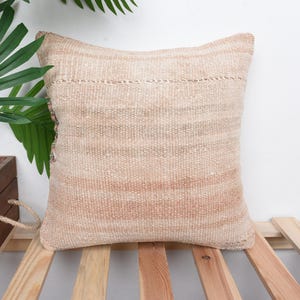 May include: A square, beige woven cushion with a textured surface. The cushion has a horizontal stripe pattern with a darker beige hue. The cushion is placed on a wooden slatted surface.
