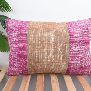 Può includere: Cuscino decorativo rettangolare con un pannello centrale marrone e pannelli laterali fucsia. Il cuscino ha un aspetto strutturato, che suggerisce un design tessuto o ricamato. È posto su una superficie a doghe di legno.