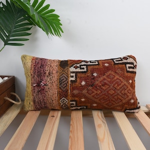 May include: Rectangular decorative pillow with a woven, textured design. The pillow features a mix of colors, including rust, yellow, and burgundy. It rests on a wooden slatted surface, with a plant in a wooden planter in the background.