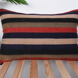 Puede incluir: Una almohada de rayas con un esquema de color beige, marrón, rojo y azul. La almohada está hecha de un tejido tejido y tiene una forma rectangular.