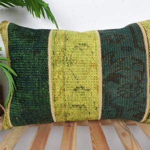 May include: Rectangular green and yellow pillow with a striped design. The pillow features a central yellow stripe flanked by darker green sections. The edges are trimmed with a lighter beige border. The pillow is placed on a wooden surface.