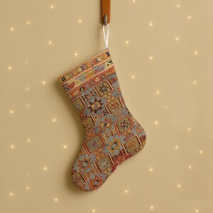 Peut inclure: Une chaussette de Noël avec un motif coloré, suspendue à un crochet en bois. La chaussette présente des formes géométriques dans des tons de bleu, orange, rouge et jaune. Un ruban blanc est attaché en haut.
