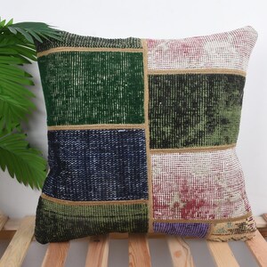 Peut inclure: Coussin carré en patchwork avec un motif géométrique. Le coussin présente une variété de couleurs, dont le vert, le bleu marine et le bordeaux. Le coussin est fait d'un tissu texturé et est placé sur une surface en bois.