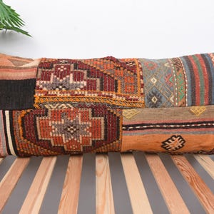 Op de afbeelding: Rechthoekig patchwork kussen met diverse geometrische patronen in oranje, rood, bruin en blauw. Het kussen ligt op een houten lattenbodem, met een groene plant op de achtergrond. De lengte is ongeveer 70 cm.