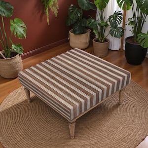 May include: Square ottoman with a brown and white striped design. The ottoman has a wooden frame and four short, light-coloured legs. It sits on a round, woven jute rug, with several potted plants in the background.