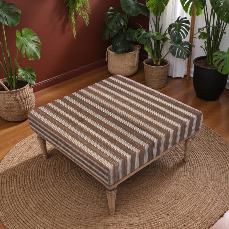 May include: Square ottoman with a brown and white striped design. The ottoman has a wooden frame and four short, light-coloured legs. It sits on a round, woven jute rug, with several potted plants in the background.