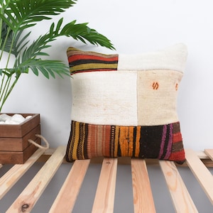 May include: A square throw pillow with a patchwork design. The pillow is made of different colored fabrics, including white, brown, orange, and red. The pillow is sitting on a wooden slatted surface.
