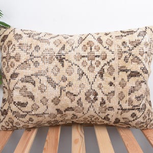 May include: Beige rectangular cushion with a distressed pattern in brown and tan. The design resembles a vintage rug, suggesting a bohemian or global style. The cushion is placed on a wooden slatted surface, possibly a bed frame.