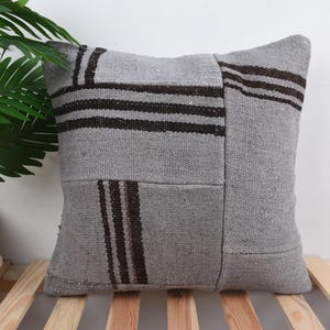 Op de afbeelding: Vierkant grijs kussen met donkerbruine strepen en rechthoekige patches. Het kussen is gemaakt van een geweven stof en ligt op een houten oppervlak. Het kussen is een decoratief item.