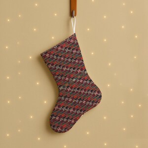 Könnte beinhalten: Ein Weihnachtsstrumpf mit einem Muster in Rot, Grün und Schwarz hängt an einem Holzhaken. Der Strumpf hat eine weiße Schlaufe zum Aufhängen und befindet sich vor einem beigefarbenen Hintergrund mit kleinen, leuchtenden Lichtern.