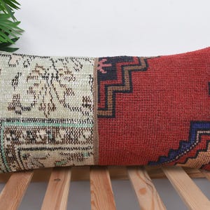 Puede incluir: Cojín decorativo rectangular con diseño patchwork. Un lado presenta una sección beige y marrón estampada, mientras que el otro es rojo liso con patrones geométricos en azul marino, marrón y dorado. El cojín está sobre una superficie de madera.