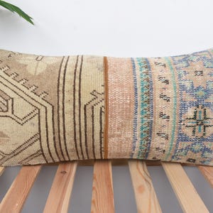May include: Rectangular decorative pillow with a patchwork design. The pillow features a beige background with geometric patterns in brown, blue, and teal. The pillow is placed on a wooden bed frame.