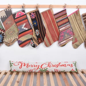 Op de afbeelding: Acht kerstsokken met diverse geweven patronen, hangend aan een houten roede. Een decoratief kussen met de tekst "Merry Christmas" ligt eronder. De sokken zijn gemaakt van stof.