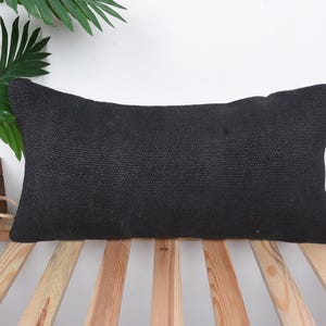 May include: A black rectangular pillow with a textured fabric surface. The pillow is sitting on a wooden slatted surface.