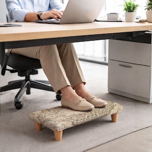 Peut inclure: Repose-pieds rectangulaire avec un dessus en tissu à motifs et quatre pieds en bois. Le tissu présente une palette de couleurs sourdes de beige, vert et marron. Le repose-pieds est placé sous un bureau, offrant un soutien ergonomique.
