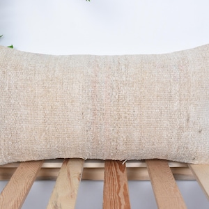 Puede incluir: Una almohada rectangular beige con textura tejida. La almohada está sentada sobre una superficie de listones de madera.