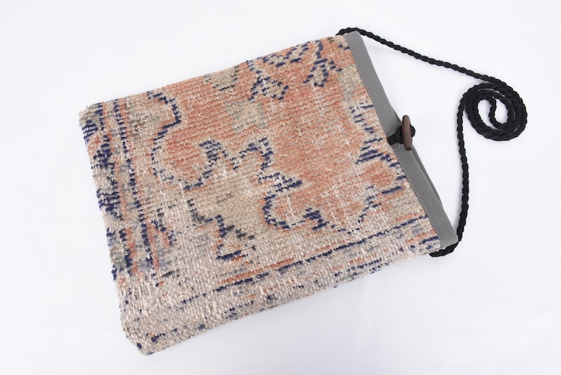 May include: A beige, blue, and orange patterned tote bag with a black adjustable strap and a wooden toggle closure.