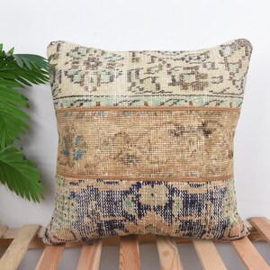 Peut inclure: Coussin carré décoratif avec un motif patchwork. Le coussin présente une section supérieure beige avec un motif floral, une section centrale beige et une section inférieure bleu foncé avec un motif similaire. Il est posé sur une surface à lattes en bois.