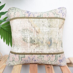 Puede incluir: Cojín decorativo cuadrado con un diseño tejido y texturizado. El cojín presenta un panel central con una mezcla de colores, incluyendo beige, verde y azul, enmarcado por bandas verde oliva. Los bordes tienen un patrón floral en rosa y morado.