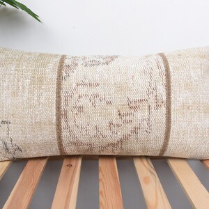 Op de afbeelding: Rechthoekig beige kussen met een geweven textuur en bruine verticale strepen. Het kussen heeft een vervaagde, vintage uitstraling, wat wijst op een traditionele of antieke stijl. Het ligt op een houten lattenoppervlak, wat bijdraagt aan de rustieke charme.