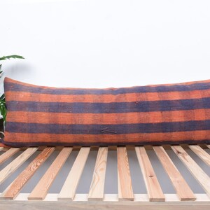 May include: A long, rectangular pillow with orange and dark blue stripes. The pillow is made of a woven fabric and is sitting on a wooden slatted bed frame.