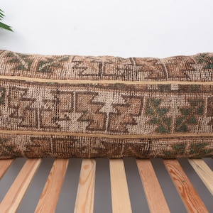 May include: Rectangular cushion with a brown and beige geometric pattern. The cushion has a woven texture and a neutral colour palette, suitable for various home decor styles. The cushion is placed on a wooden bed frame.