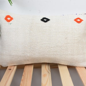 Puede incluir: Una almohada rectangular blanca con diseños bordados en naranja y negro. La almohada está sobre una superficie de madera.