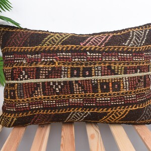May include: Rectangular decorative pillow with a woven, geometric pattern in shades of brown, red, yellow, and white. The pillow has a textured appearance and is placed on a wooden slatted surface. The pillow is a home decor item.