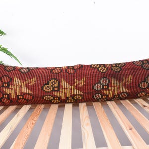 May include: A long, rectangular pillow with a red background and a repeating pattern of yellow and black shapes. The pillow is placed on a wooden slatted surface, with a green plant in the background.