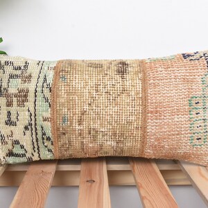 May include: Rectangular decorative pillow with a patchwork design. The pillow features sections of beige, brown, and peach tones with intricate patterns. The pillow is placed on a wooden slatted surface.