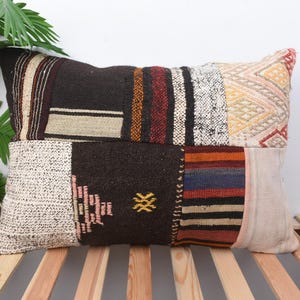 May include: Rectangular patchwork pillow with a variety of textures and colors. The pillow features sections of dark brown, cream, and patterned fabrics with accents of red, orange, and yellow. The pillow is placed on a wooden slatted surface.