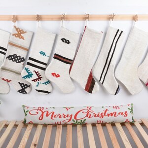 Pode incluir: Nove meias de Natal brancas com padrões coloridos tecidos pendurados em um trilho de madeira. Uma almofada branca com o texto "Merry Christmas" em vermelho está em um banco de madeira.
