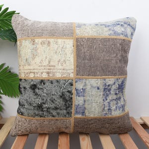 May include: Square patchwork cushion with a geometric design. The cushion features beige, brown, black, and blue panels. It is made of woven material and has a textured appearance. The cushion is approximately 40cm x 40cm.