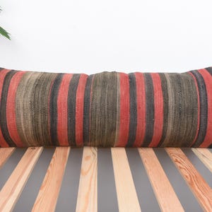 Puede incluir: Un cojín rectangular y alargado con un diseño a rayas en tonos rojos, negros y marrones. Descansa sobre una superficie de listones de madera. El patrón y los colores del cojín crean una estética cálida y acogedora, ideal para una cama o banco.