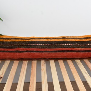 May include: A long, rectangular pillow with a striped pattern in shades of orange, red, and brown. The pillow is made of woven fabric and has a rustic, handmade look.