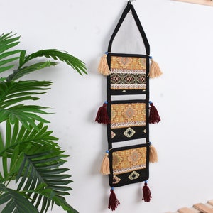Può includere: Un organizzatore da parete con tre tasche, ognuna con un diverso motivo geometrico in tonalità di giallo, nero e marrone. Le tasche hanno nappine rosse, crema e blu.