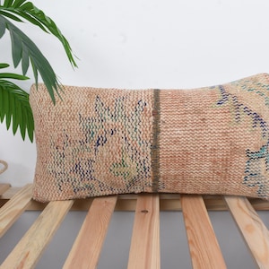 May include: A rectangular cushion in beige with a woven texture and faded blue and green patterns. The cushion is on a wooden slatted surface. A green plant and wooden crate are in the background.