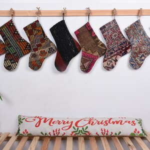 May include: Eight colorful Christmas stockings made from repurposed rugs hang on a wooden rod. The stockings are decorated with various patterns and colors, including red, green, blue, and brown. A pillow with the text "Merry Christmas" is on a wooden bench below the stockings.