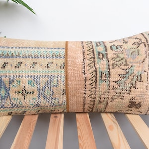 May include: Rectangular beige pillow with a vintage rug design. The pillow features a patchwork design with sections of blue, teal, and brown patterns. The pillow is placed on a wooden slatted surface.