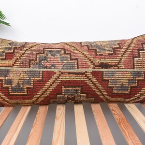 May include: Rectangular pillow with a geometric pattern in shades of red, brown, and beige. The pillow is placed on a wooden bed frame, with a green plant in the background.
