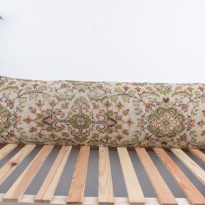 May include: Long, rectangular pillow with a beige background and an intricate floral pattern in shades of green, brown, and orange. The pillow is placed on a wooden slatted surface, suggesting a bed or bench.