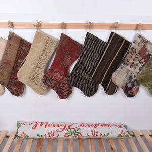 Peut inclure: Neuf chaussettes de Noël colorées faites de tapis recyclés sont accrochées à un support en bois. Les chaussettes sont décorées de motifs et de couleurs variés, notamment le rouge, le vert, le marron et le beige. Les chaussettes sont accrochées à un support en bois sur un fond blanc.