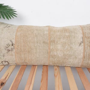 May include: Rectangular beige pillow with a textured surface and vertical stripes. The pillow is placed on a wooden bed frame, with a green plant and a wooden box in the background.