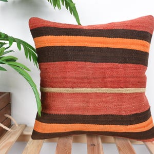 May include: Square decorative pillow with a striped pattern in shades of orange, brown, and red. The pillow is made of woven fabric and is placed on a wooden surface. A plant is visible in the background.