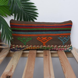 May include: A rectangular brown woven pillow with colorful horizontal stripes and tree-like designs. The pillow has a brown background with orange, green, and blue tree designs. The pillow is placed on a wooden bed frame.